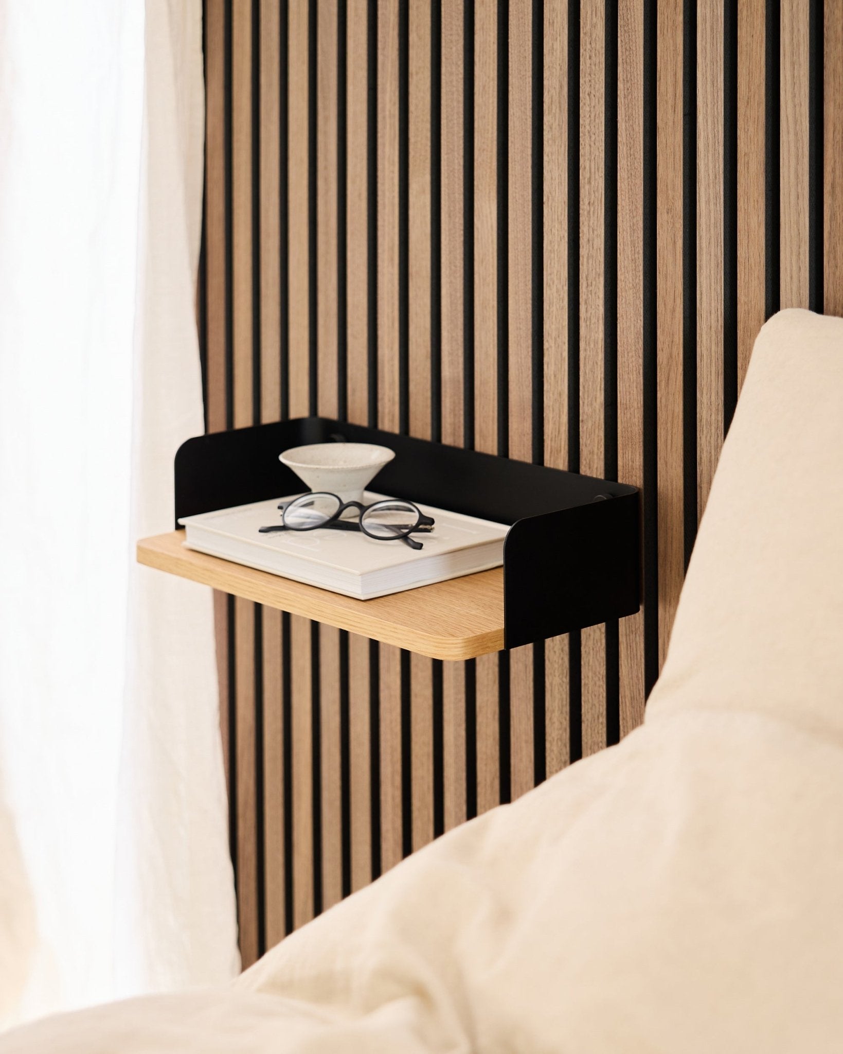 The Linn Bookshelf by WoodUpp in natural oak (39.9 cm) with a book, eyeglasses, and a glass beside a bed against an Akupanel acoustic wood panel wall.